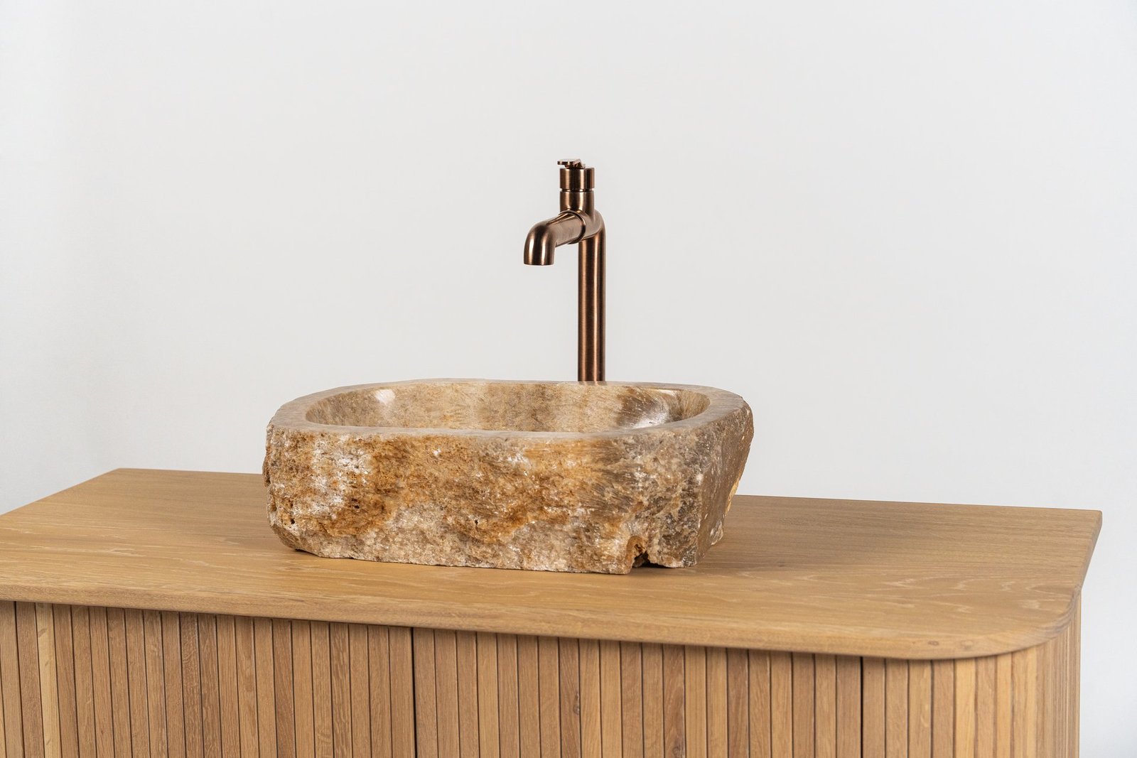 Lavabo rectangular de piedra natural rústica con grifo de bronce, sobre mueble de baño de madera clara.